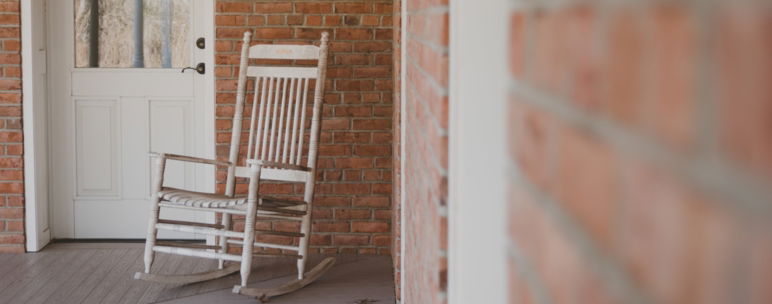 White, wooden rocking chair on a wooden porch in front of a brick building.